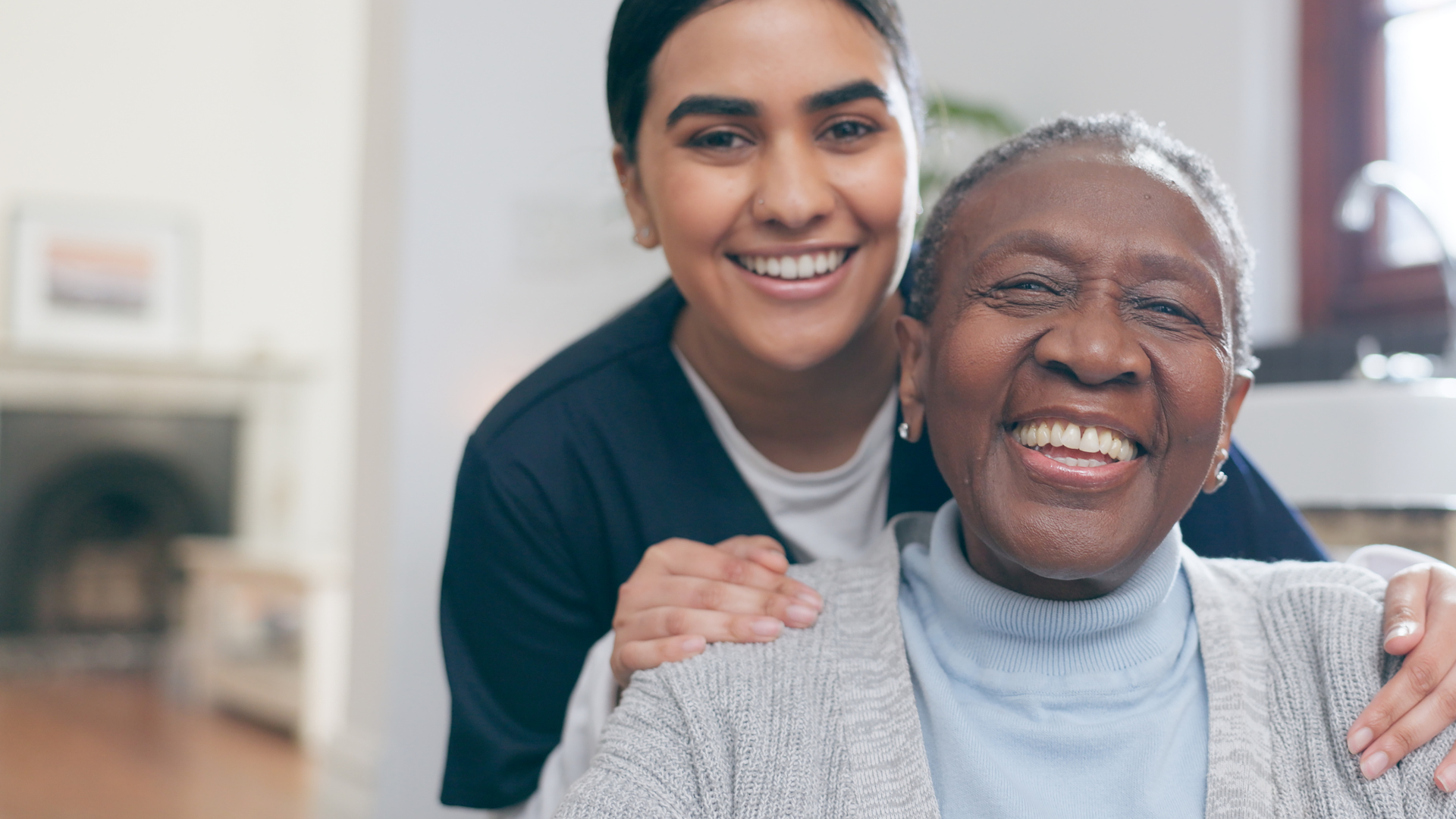 Smiling caregiver with her hands on an older adult's shoulders in a bright home